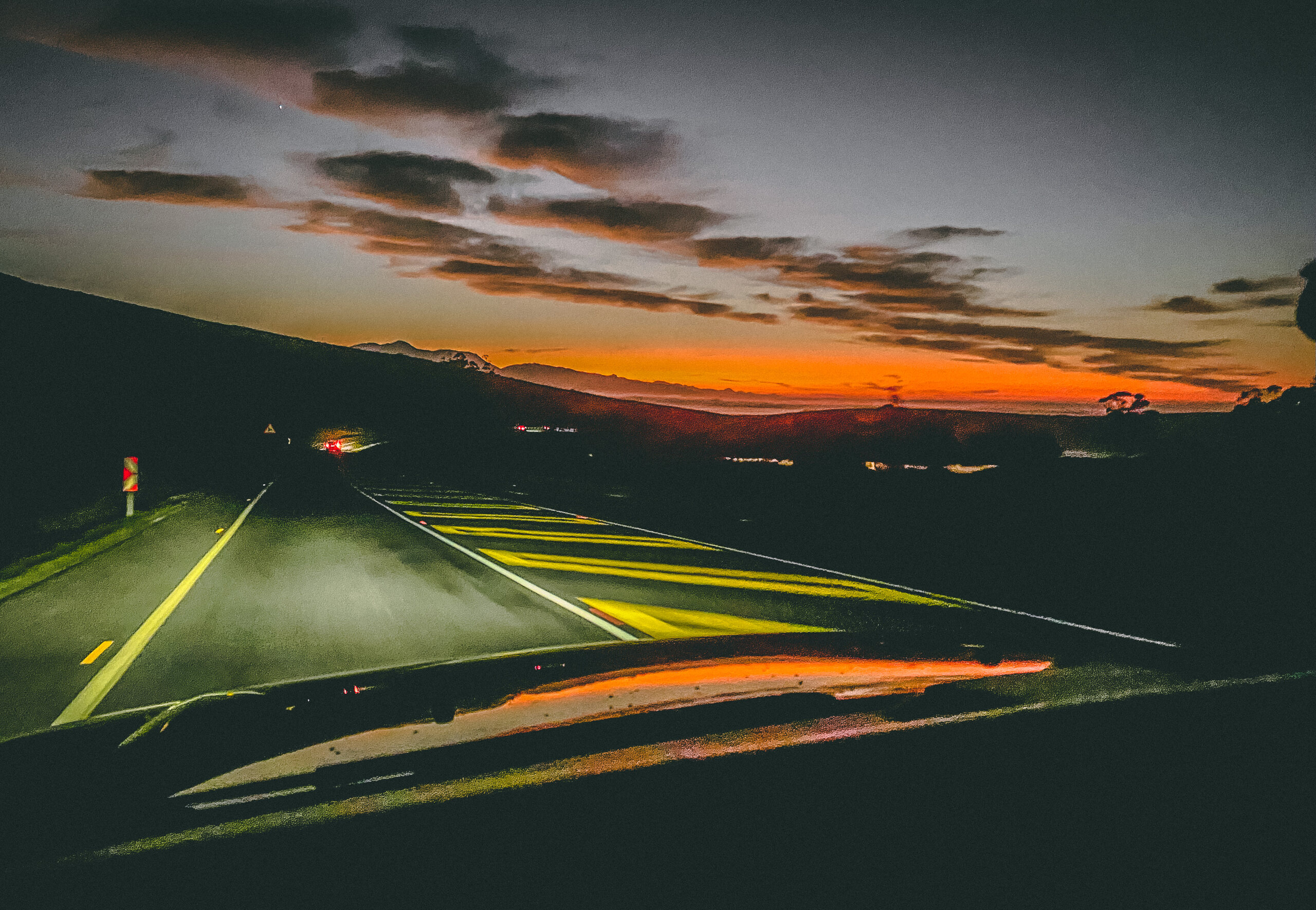 A road leading towards traffic and a red horizon beneath evening clouds.